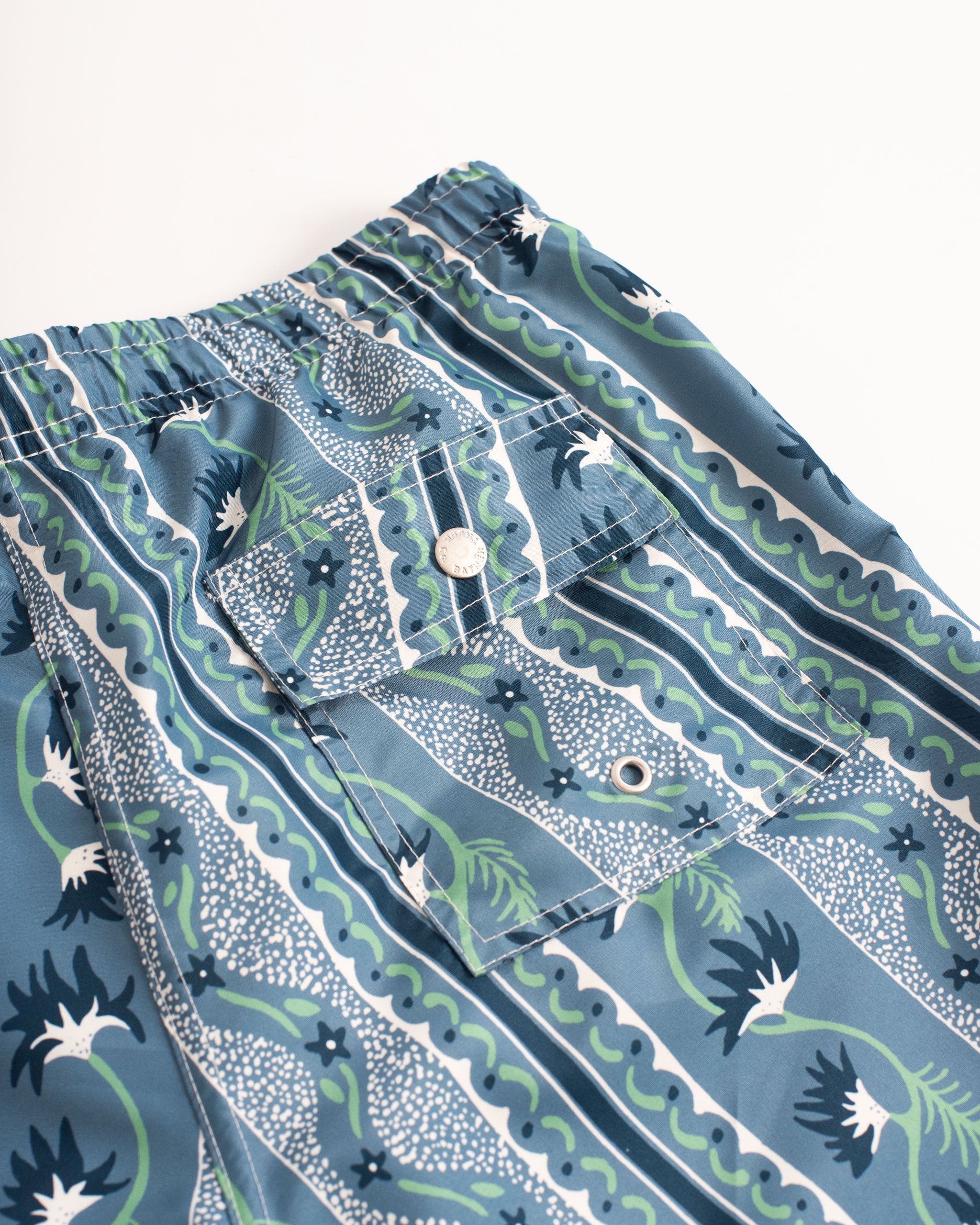 back pocket close up of Blue swim trunk with classic stripe features intricate mosaics of flowers, sand, and starfish