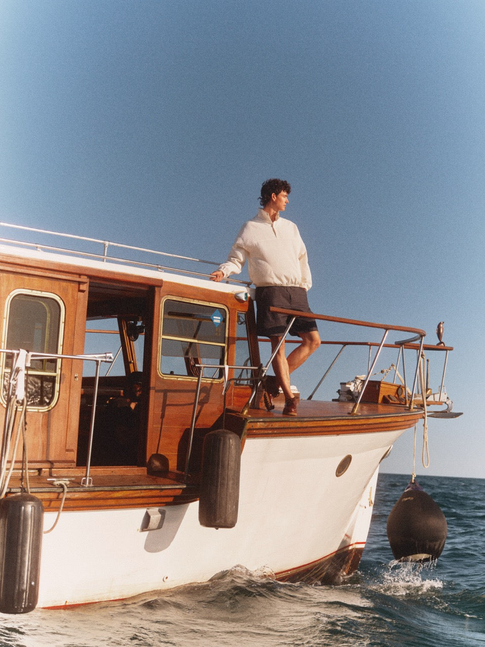 Man standing on a vintage boat looking out to the sea in a white pullover and navy shorts.