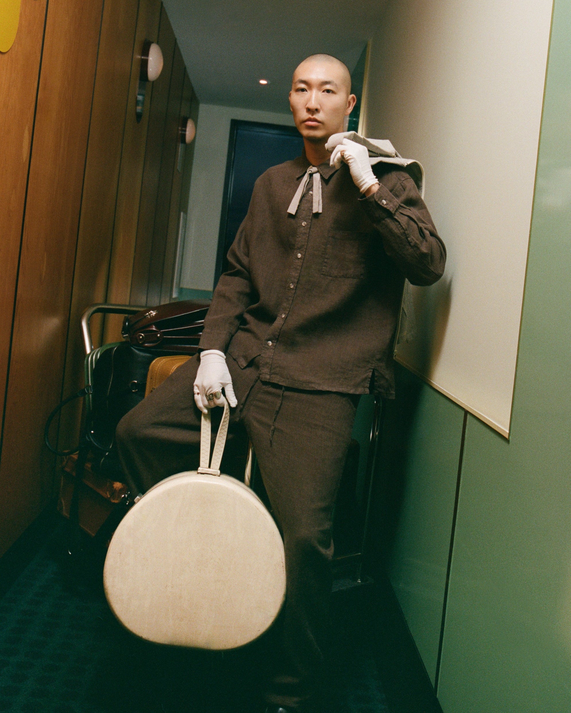 Man in a brown outfit holding a round beige bag in a hallway.