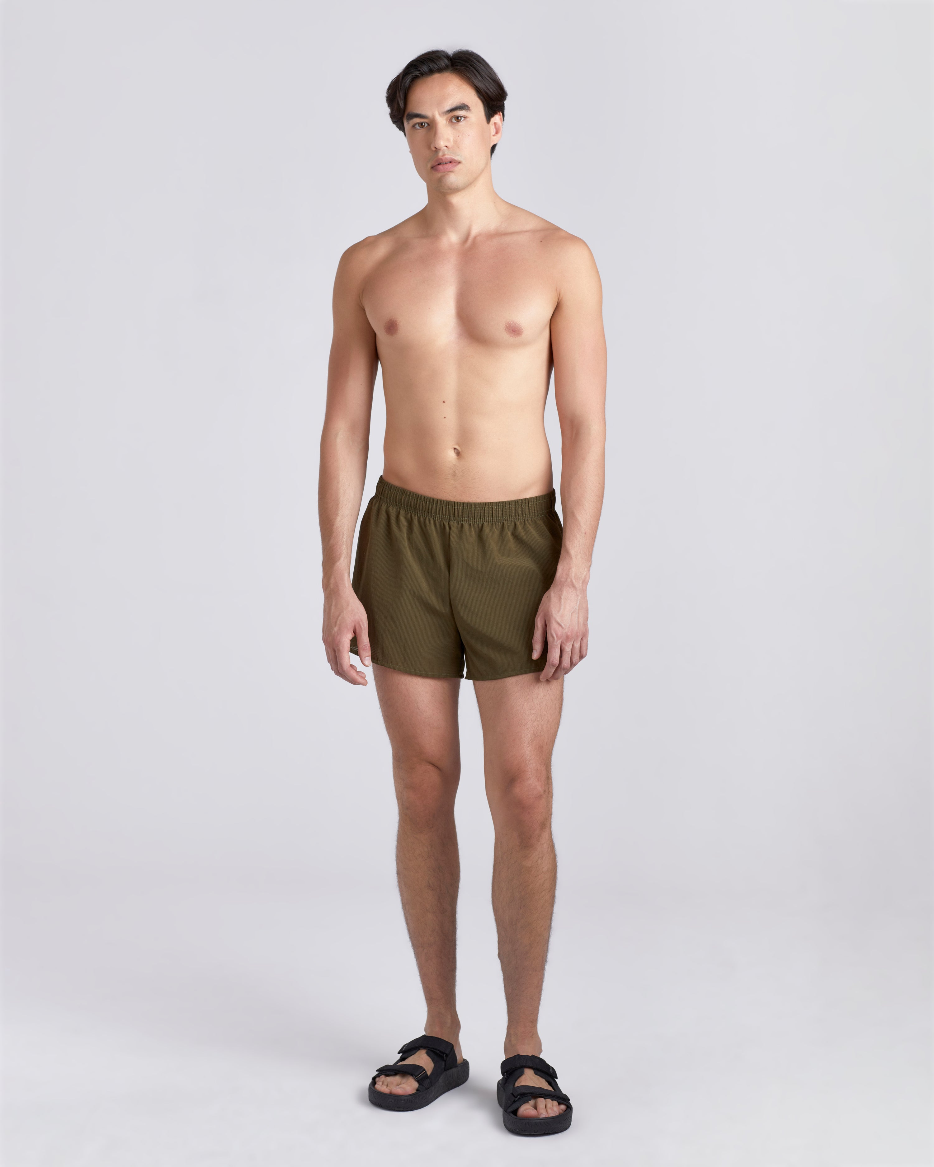 Front view of shirtless model wearing sandals and 3.5 inch inseam Bather Active Trunk in green featuring lightweight recycled Japanese DotAir nylon with stretch fabric and compression liner