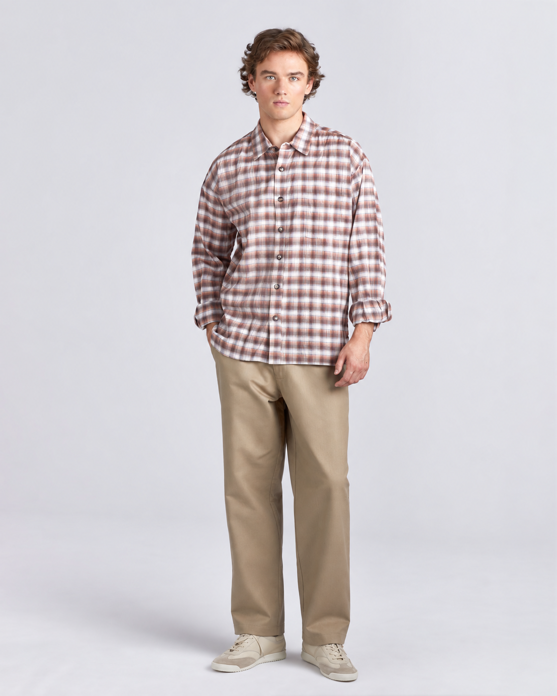 A young man modeling a brown and white checkered long-sleeve button-down shirt, paired with beige trousers and light beige sneakers, against a light gray studio background.