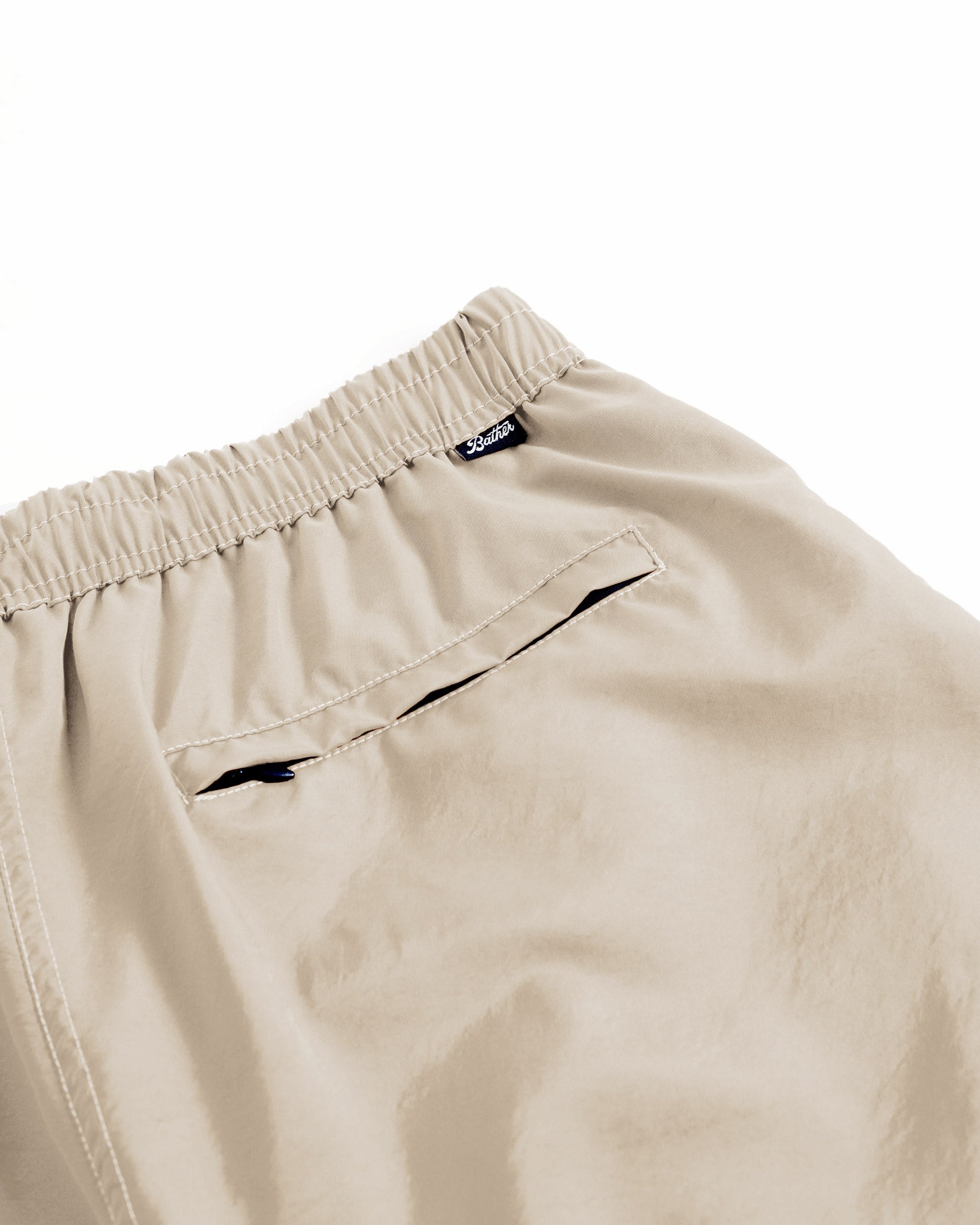 Shiny beige swim trunks with a brand Bather label and zippered pocket on a white background