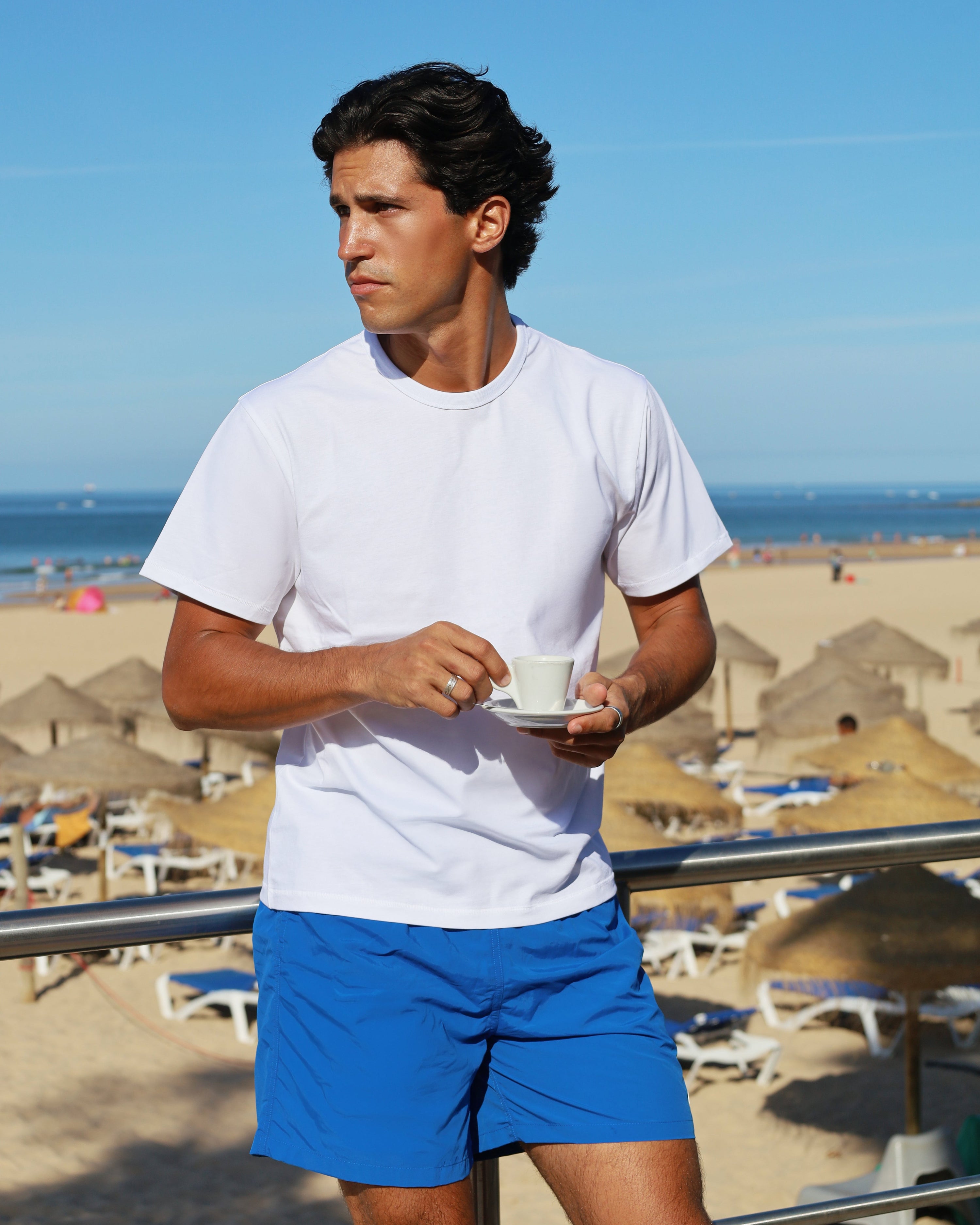 A man standing on a beach balcony wearing a white crewneck t-shirt and blue athletic shorts, holding a white coffee cup and saucer.
